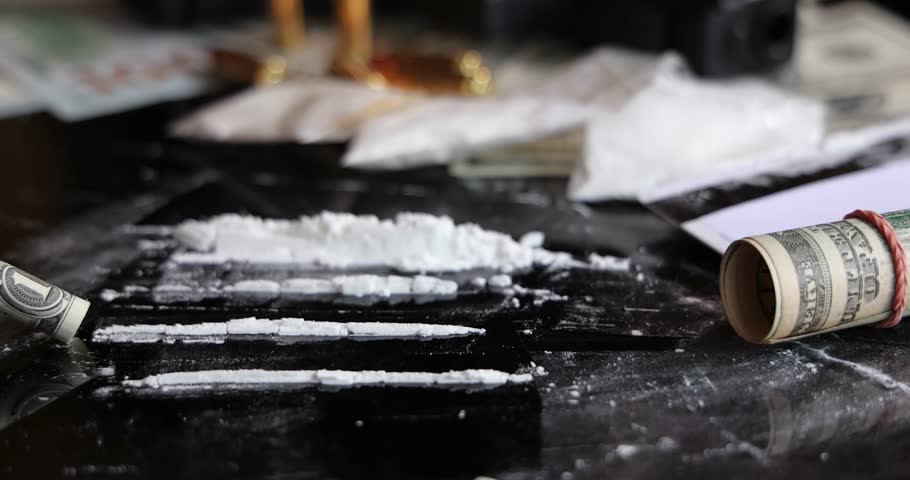 Snorting a line of cocaine with a rolled dollar bill on black background, slow motion. Illegal drugs and cash with gun and bullets on table - crime scene. Criminal problems. Illegal selling