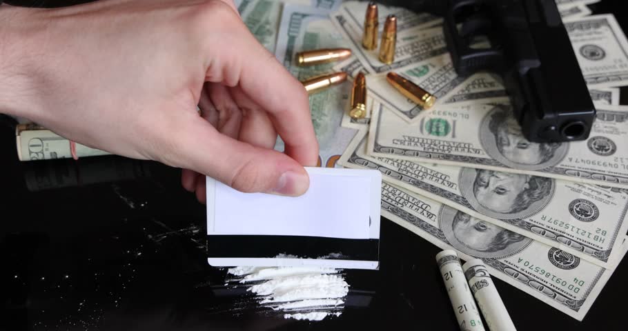 Illegal drugs and cash with gun and bullets on table - crime scene. Man making cocaine line with credit card. Criminal problems. Drugs and money on black background. Illegal selling