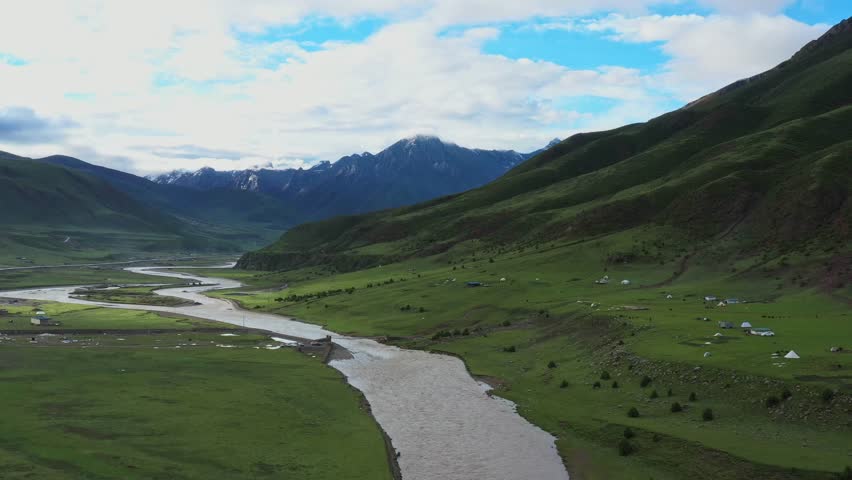 A majestic river carves its path through a verdant valley, flanked by gentle slopes and distant, imposing mountains under a dynamic sky.
