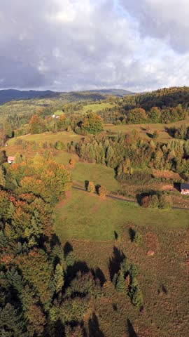 Aerial view of autumn countryside with green fields, colorful trees, and mountain hills under moody cloudy sky, Scenic landscape showing the beauty of fall season in the mountains.