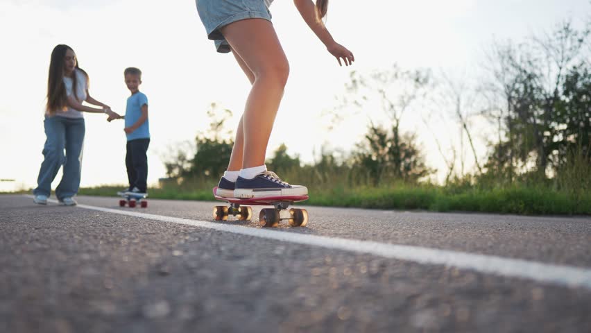 A girl on a skateboard glides down the road while another girl follows on the road. The boy stands on his skateboard, observing the road and interacting with the girl.