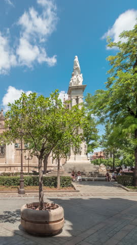 Plaza del Triunfo timelapse hyperlapse with Monument to Immaculate Conception and Cathedral of Seville. Green trees, historic architecture, blue sky with clouds. Scenic city view in Andalusia, Spain