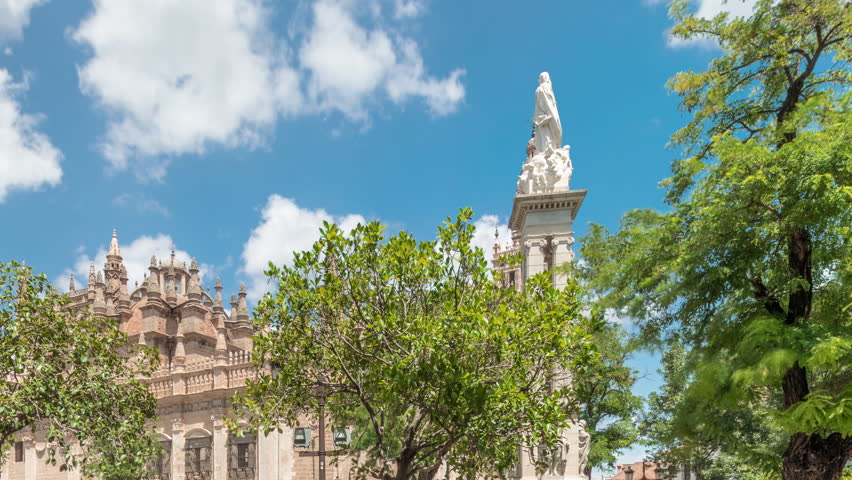 Plaza del Triunfo timelapse hyperlapse with Monument to Immaculate Conception and Cathedral of Seville. Green trees, historic architecture, blue sky with clouds. Scenic city view in Andalusia, Spain