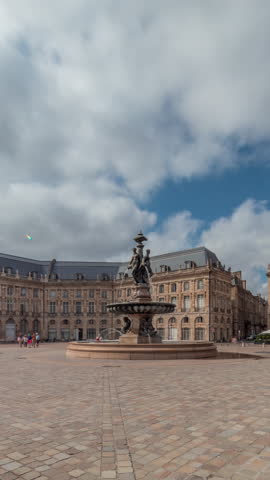 Place de la Bourse timelapse hyperlapse in Bordeaux, France, with the Fountain of the Three Graces surrounded by classic architecture under a cloudy blue sky. People walking around the main square