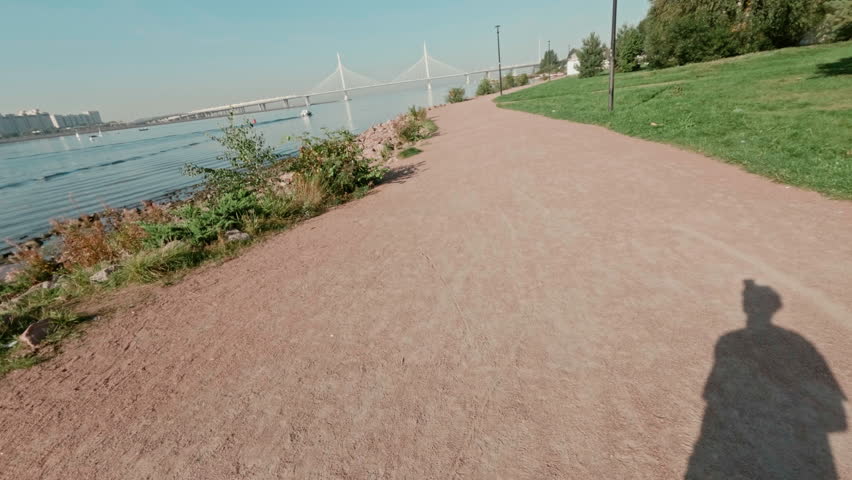 POV of sportsman with action camera capturing outdoor jogging session along long sandy road between green lawns and trees growing in park and riverside