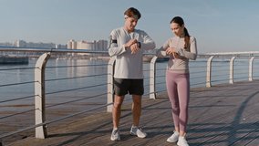 Push in of young male and female athletes in activewear standing on wooden deck by riverside, checking time on their smartwatches, looking at one another and starting morning run - Powered by Shutterstock - Get 15% off with code: PIKWIZARD15