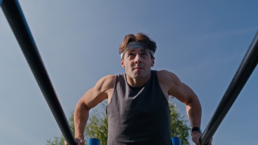 Young male athlete wearing grey t-shirt and headband performing dips on parallel bars of sports facilities, then stopping doing exercise and having short break