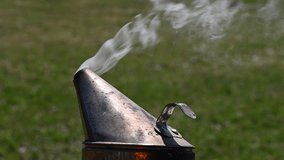 A close up of the spout of an old beekeeping smoker with smoke coming out, green grass in the background - Powered by Shutterstock - Get 15% off with code: PIKWIZARD15