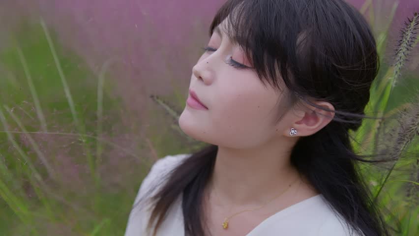 A beautiful Asian girl sits among pink muhly grass, closing her eyes to feel the gentle breeze on her face.