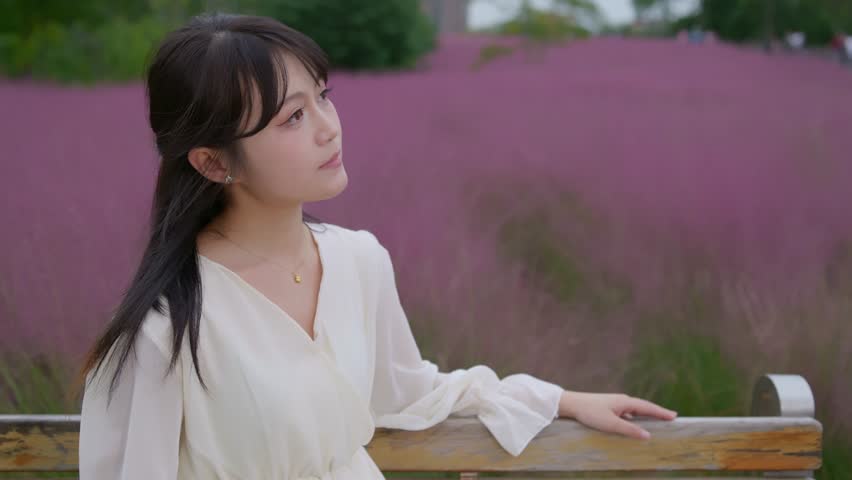 A beautiful Asian girl wearing a white long dress is among pink muhly grass, with the camera moving from a distance to close-up on her beautiful face.