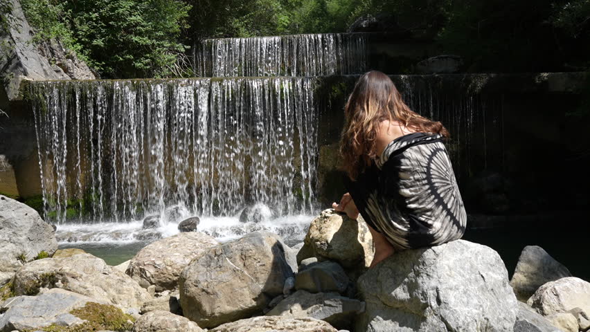 Back view of woman sitting on rocks watching a small waterfall in Swiss nature near Walensee