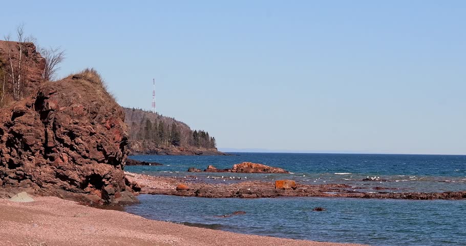 Waves roll onto a beach at the rocky shore of Lake Superior, as seagulls fly over and float on the blue lake water, on a sunny day in Minnesota.