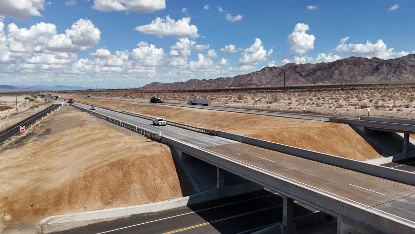 Drone View of Highway in Desert, Static Shot of Interstate in Desert Center California, Mountain range in background, Bright Blue Sky and white clouds, cars and semis driving on road