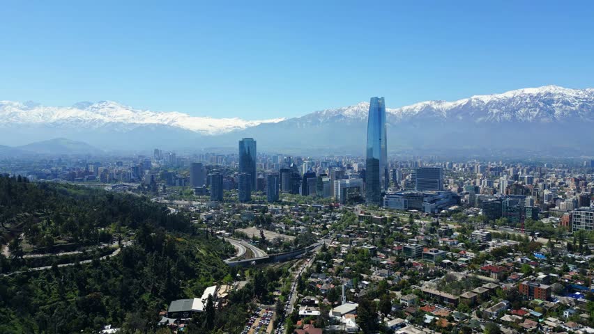 Drone aerial descending past the glass Costanera tower, revealing downtown skyline and snowcapped Andes under clear daytime light. Santiago, Chile