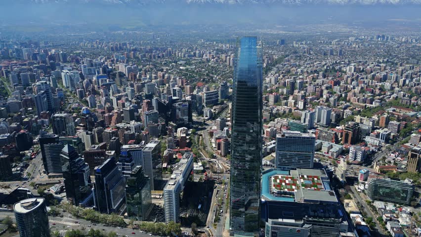 Drone aerial orbit revealing Costanera skyscrapers, highways and snowcapped Andes under clear daytime skies. Santiago, Chile