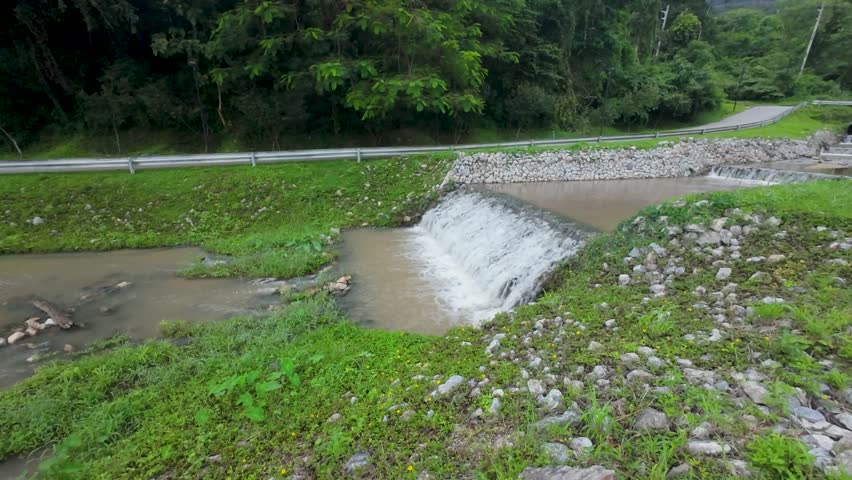 Scenic view of a small, cascading waterfall in a muddy river next to a winding road and lush green forest.