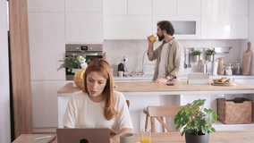 Young businesswoman working on her laptop in the kitchen and talking on the phone while her husband drinks juice in the background, showing a modern family lifestyle with remote work balance - Powered by Shutterstock - Get 15% off with code: PIKWIZARD15
