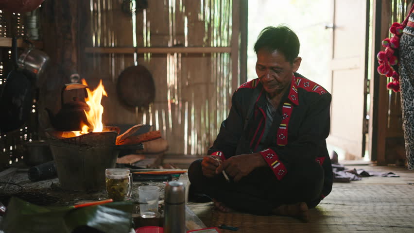 Elderly aismatic man sitting on the ground barefoot lighting a traditional craft cigarette while sitting inside a wooden dwelling