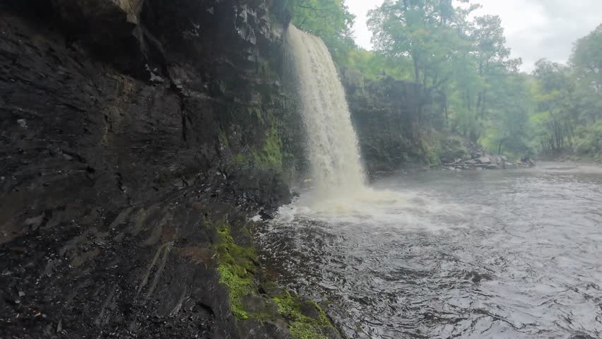 A scenic drone video captures the tranquil Angel Falls in Brecon Beacons, surrounded by lush green foliage and rugged rocks. A person in a jacket stands in awe near the serene cascade.