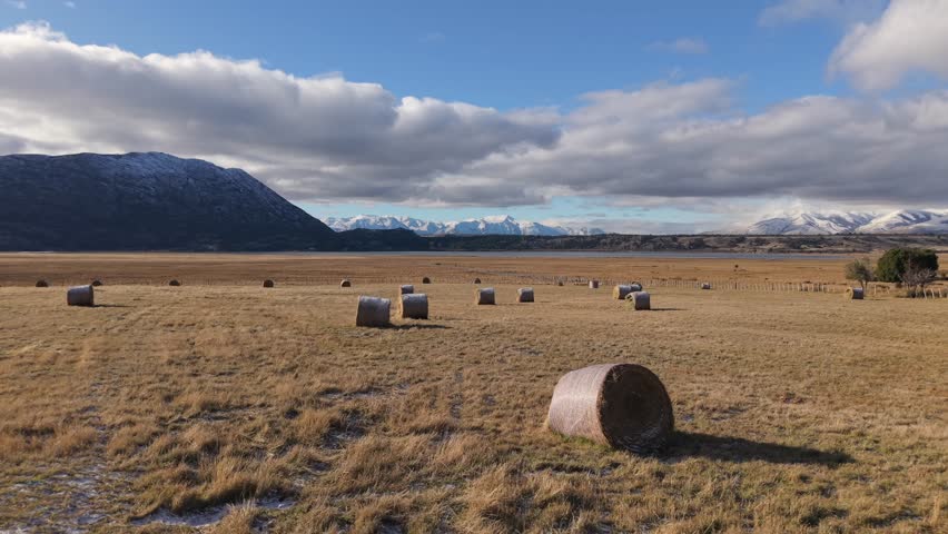 Drone pulls back over golden hay bales scattered across the Patagonian steppe—revealing the vast field beneath snow-capped Andes peaks in Chubut, Argentina, revealing farming and livestock tradition