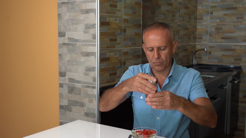 A 60-year-old Man Eating a Mixed Fresh Salad at his Home. A Middle-aged man in a blue short-sleeved shirt sitting at the dining table, enjoying a mixed vegetable salad dressed with olive oil.