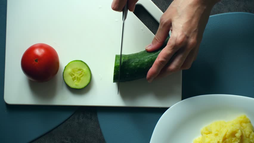 Chef slicing fresh cucumber, tomato on cutting board, then plating them with mashed potatoes, grilled salmon steak for delicious, healthy home cooked meal. Slicing vegetables for healthy fish dinner
