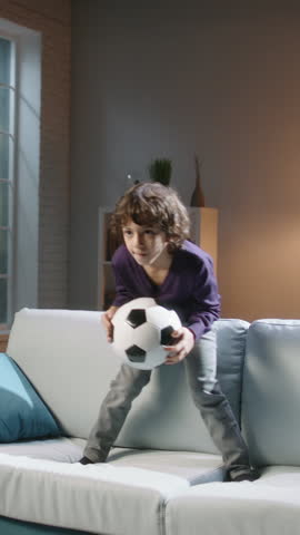 Excited Boy Jumping on Sofa with Soccer Ball, Watching Football Match on TV, Cheerful Child Celebrating Goal, Home Sport Fun, Family Leisure, Happy Childhood, Energy and Joyful Emotions