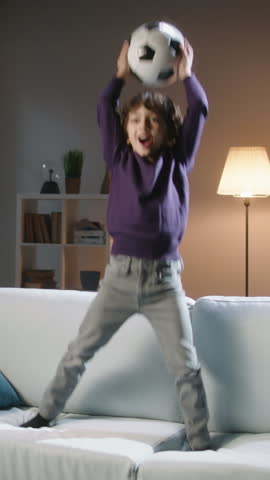 Excited Boy Jumping on Sofa with Soccer Ball, Watching Football Match on TV, Cheerful Child Celebrating Goal, Home Sport Fun, Family Leisure, Happy Childhood, Energy and Joyful Emotions