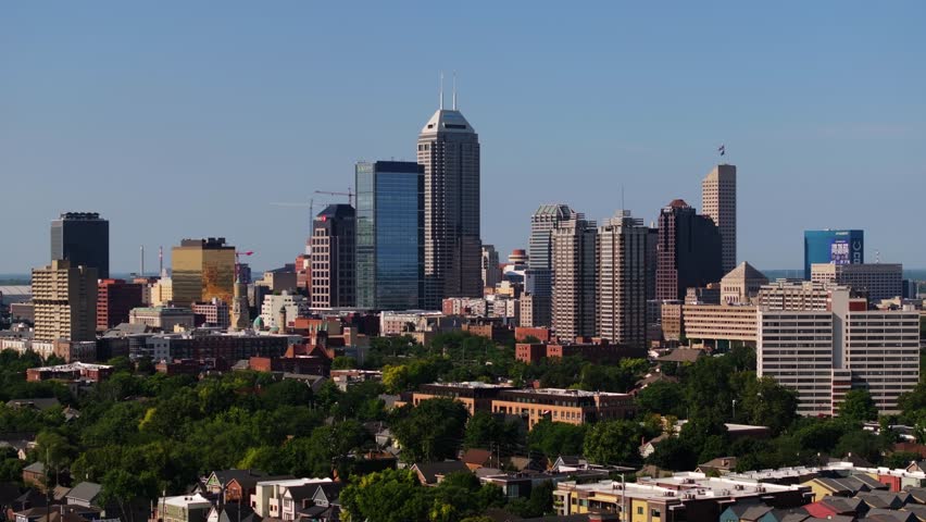 Drone slides sideways over green park revealing downtown Indianapolis skyline on sunny day.