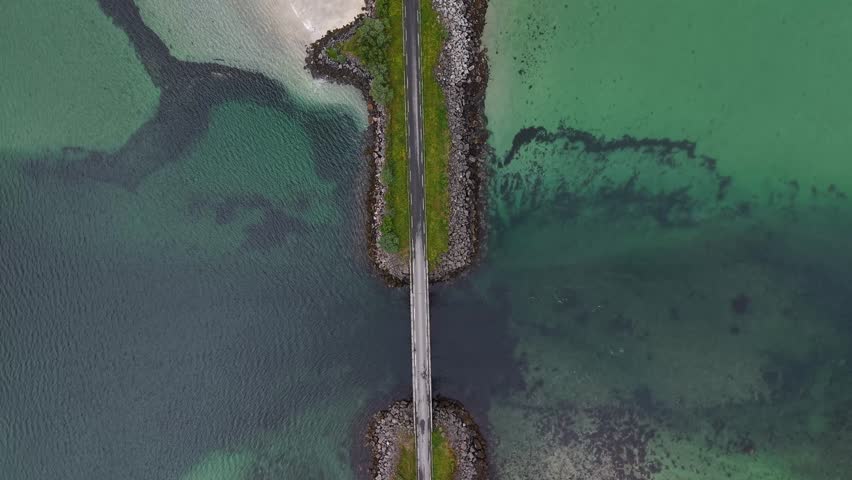 Drone view of road running between two islands in Arctic Lofoten, Norway, surrounded by vivid turquoise fjord water and sandbanks.