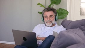 Happy senior man with a beard wearing headphones and talking on a video call on his laptop, gesturing and smiling while sitting comfortably on the sofa in his living room - Powered by Shutterstock - Get 15% off with code: PIKWIZARD15