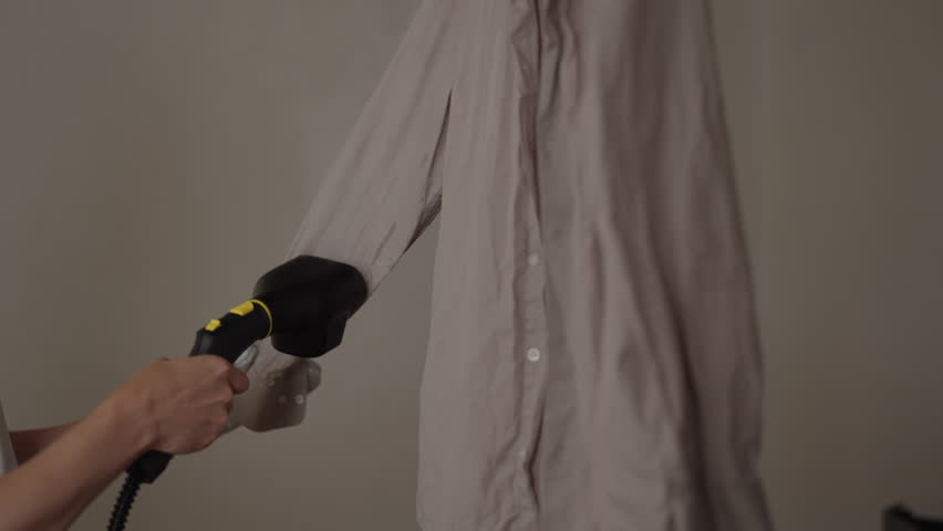 Unrecognizable person using handheld clothes steamer, removing wrinkles from beige collared shirt, simplifying household chores and garment care with modern ironing alternative technology, close-up.