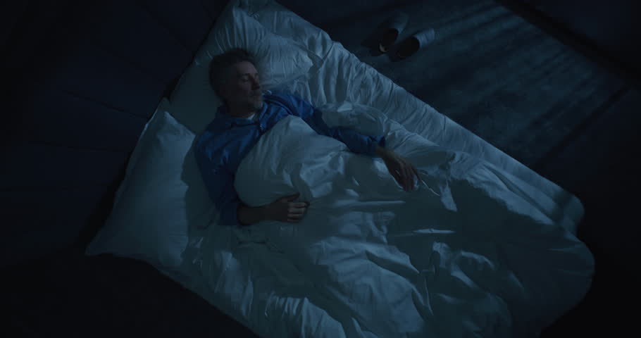 Exhausted by Insomnia, Mature Man Irritably Gets Out of Bed in Bedroom Lit By Moonlight. Tired and Upset, Puts on Slippers and Leaves Room. Poor Sleep, Frequent Nighttime Awakening. Rotating Top View.