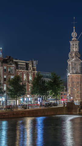 Panoramic timelapse of Amsterdam city center featuring Amstel River, waterfront and Munttoren tower. Boats float back and forth at night. Scenic urban view from bridge. Netherlands