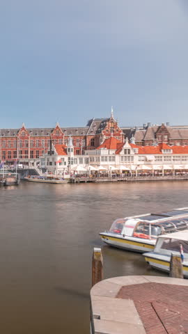 Timelapse of Amsterdam Central Railway Station front view with tour boats moored at the jetty, trams passing, and a canal in the foreground. Amsterdam, The Netherlands