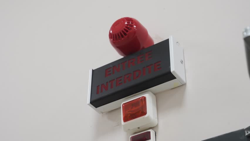 A close-up on a wall-mounted warning system for a restricted area. A sign reads "ENTREE INTERDITE" (Do Not Enter) and is fitted with an alarm siren and flashing light.