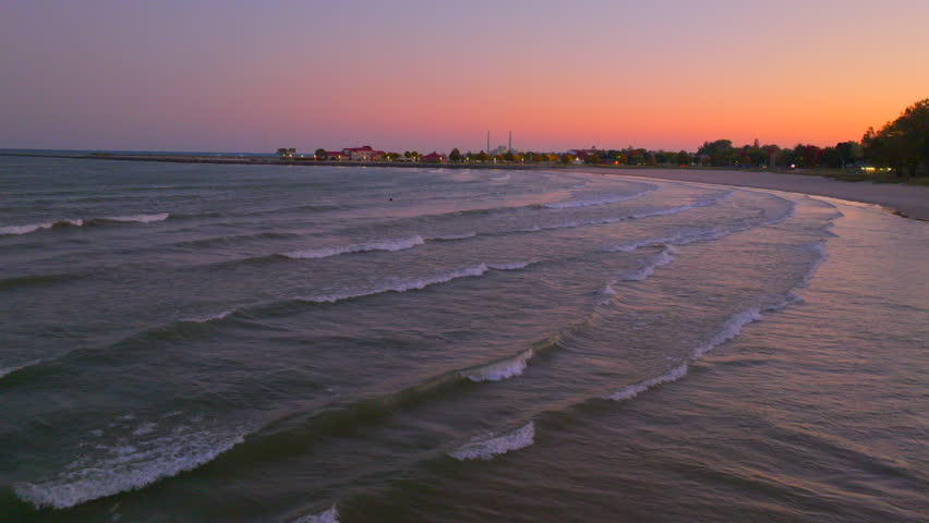 Drone aerial at sunset over Lake Michigan in Sheboygan, Wisconsin, pulling back across gentle waves, concrete piers, and sandy beach under warm evening light.