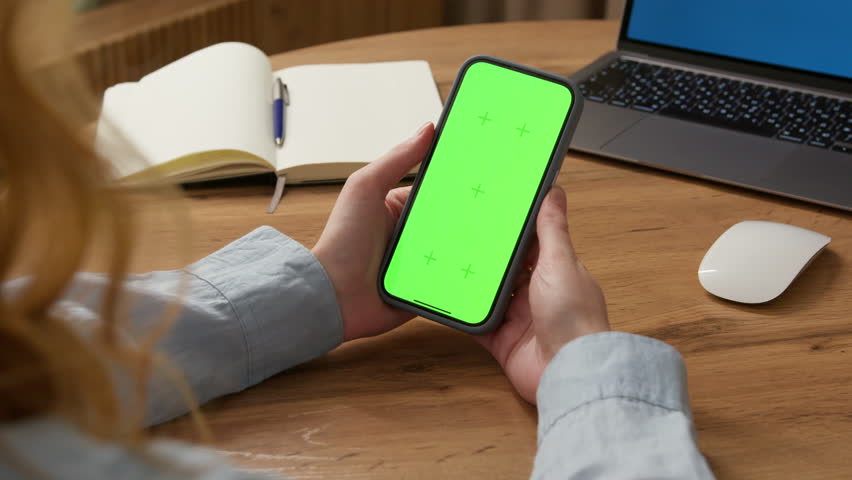 Woman sit use digital tech cell phone, green screen mock up. Hand scroll iphone web site, swipe like photos. Girl hold mobile closeup. Chroma key page. Cozy office background. Online app copy space.