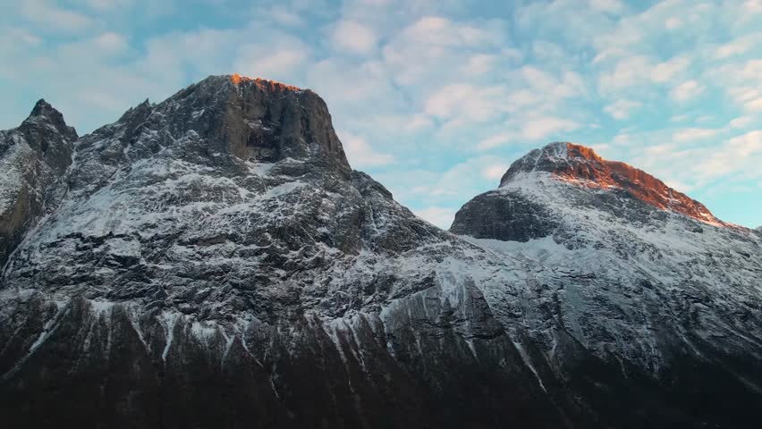 Aerial view of the iconic Trollstigen mountain road winding through snow covered peaks during a vibrant sunset, showcasing the dramatic Norwegian landscape and its unique natural beauty. Perfect for t