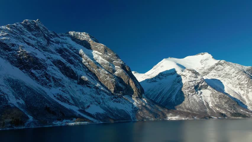 This drone footage capturing the serene beauty of Eresfjorden in winter, showcasing majestic snow covered mountains towering over calm, reflective waters under a clear blue sky.