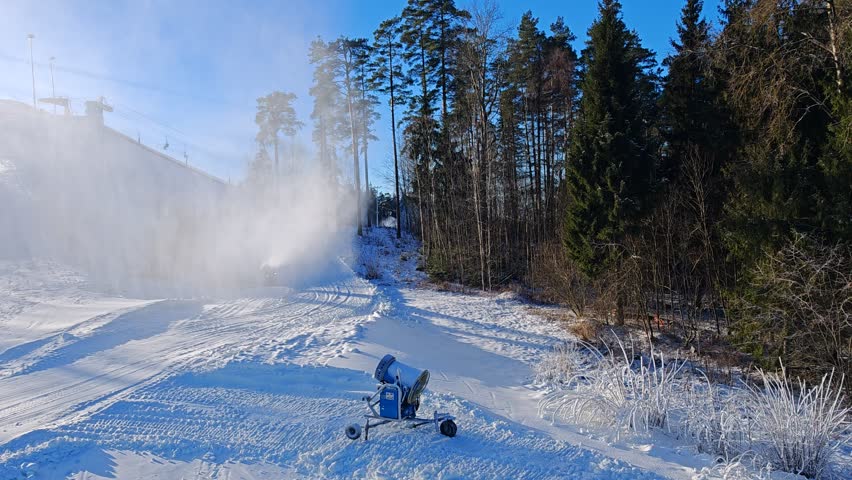 There are chairlifts on the slopes of the ski center. Skiers climb them. Snow cannons are working. Mesh fences are installed for safety. There are footprints in the snow. Nearby bushes and pines