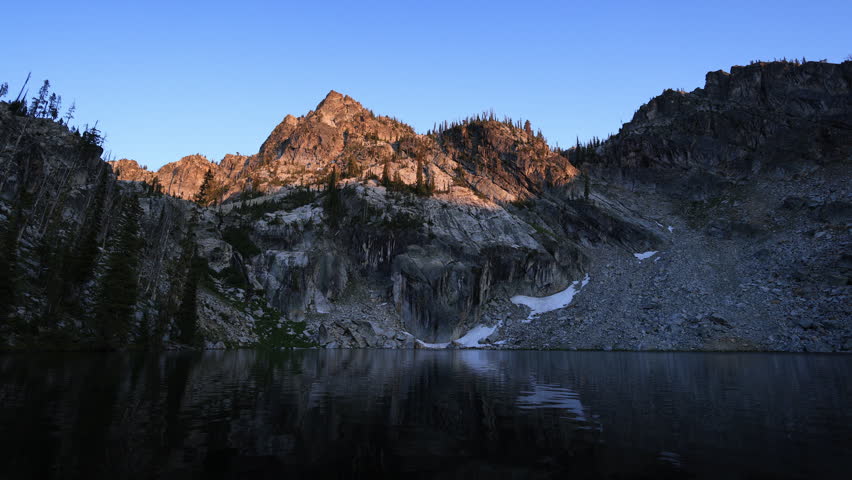 Sunlight illuminates the rugged peaks above Lower Trail Creek Lake in Idaho’s Sawtooth National Forest at sunset, above calm alpine waters.