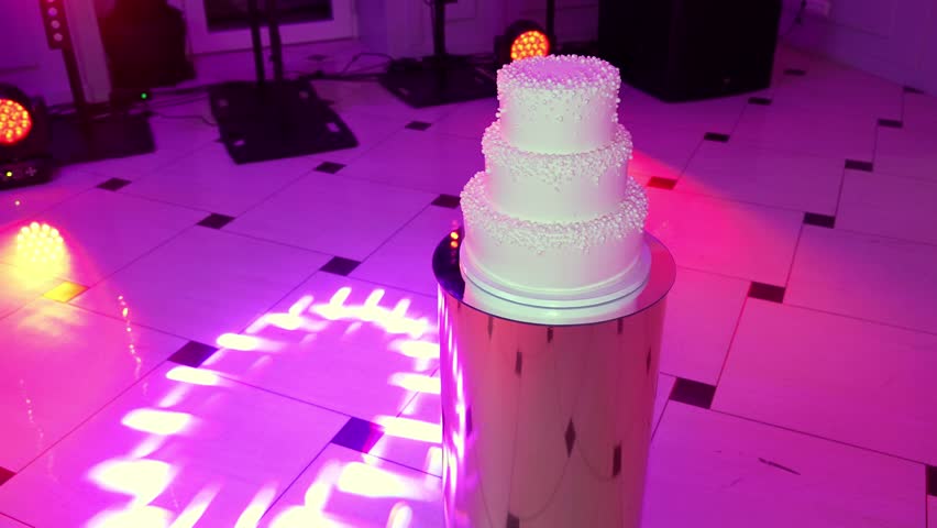 A scenic view of a three-tier white cream cake placed on a stand in a lighted hall