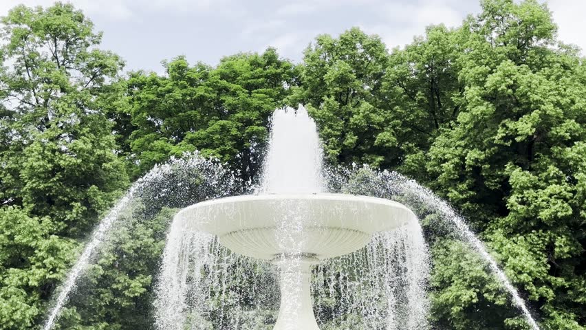Elegant white fountain spraying water upwards in a lush park setting, surrounded by dense green trees on a bright summer day.