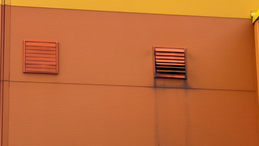Bright orange industrial wall features two vents; right vent moves up and down creating subtle rhythmic motion, evoking urban atmosphere, energy, and industrial character.
