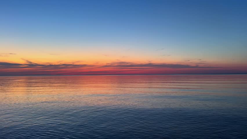 Peaceful sunset over quiet ocean paints blue and orange tones across water. Gentle waves reflect vivid colors creating calm mood and wide open horizon.