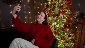 Woman in red sweater and reindeer headband makes a video call on smartphone in front of a glowing Christmas tree, sharing festive greetings and holiday joy perfect for seasonal design themes - Powered by Shutterstock - Get 15% off with code: PIKWIZARD15