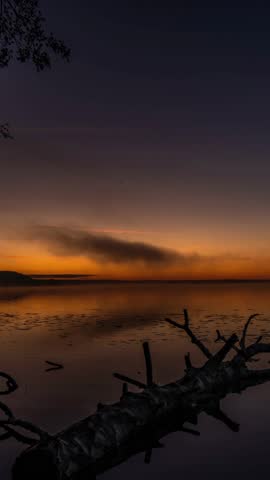 A tranquil sunset scene unfolds over a calm lake, featuring a vibrant orange and purple sky reflected in the still waters.