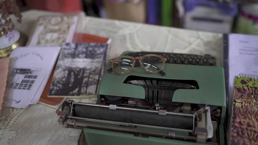 Close-up of a vintage green typewriter with eyeglasses resting on top, surrounded by papers and notebooks on a lace-covered table, evoking a nostalgic writing atmosphere - parallax shot