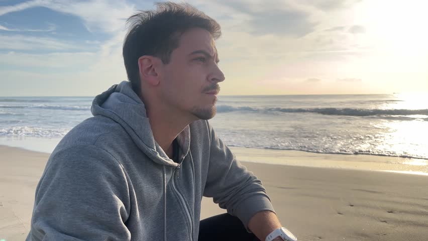 A man is sitting on the beach with his head down. He is wearing a gray hoodie and a black shirt. The sky is cloudy and the sun is setting